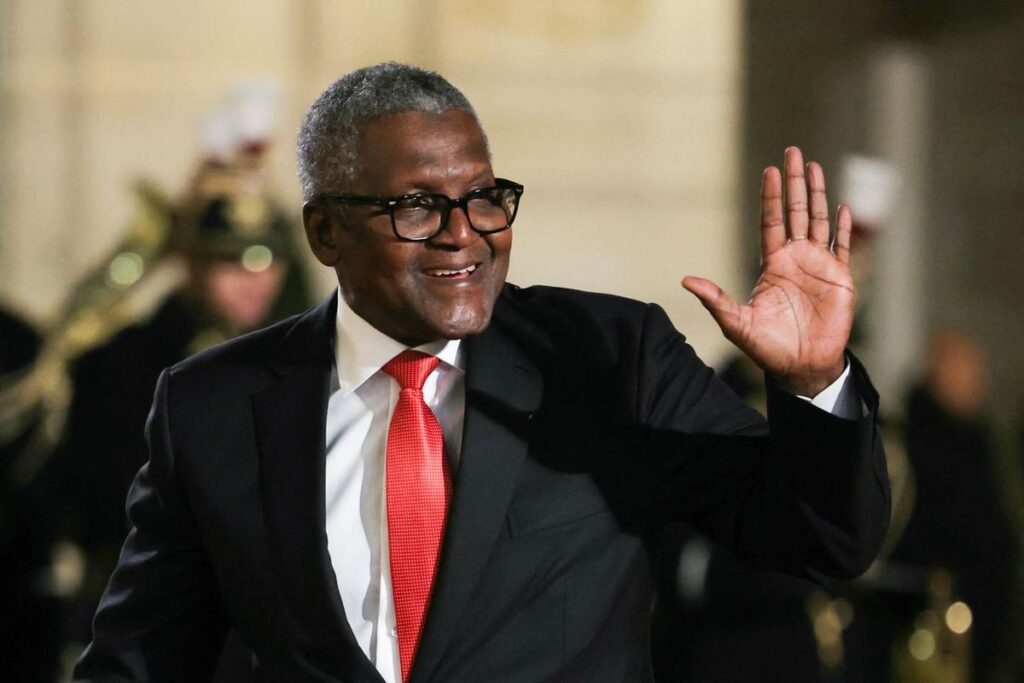 An image of Aliko Dangote in a black suit with a white shirt and red tie, waving and smiling
