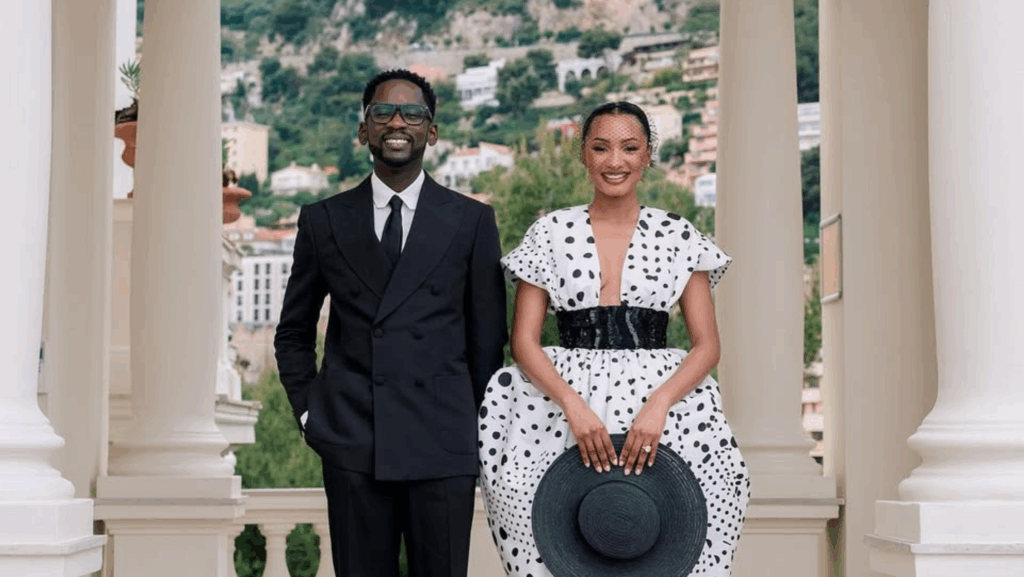 Mr Eazi and Temi Otedola pose together in elegant outfits at their Monaco wedding, with scenic hills and architecture in the background. Mr Eazi wears a black suit, while Temi stuns in a white polka dot dress with a black belt and matching hat.