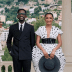 Mr Eazi and Temi Otedola pose together in elegant outfits at their Monaco wedding, with scenic hills and architecture in the background. Mr Eazi wears a black suit, while Temi stuns in a white polka dot dress with a black belt and matching hat.