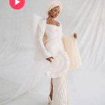 Temi Ajibade dressed in a fitted off-white gown with dramatic sleeves and a gele headwrap holds a matching hand fan while posing in front of a soft white draped background.
