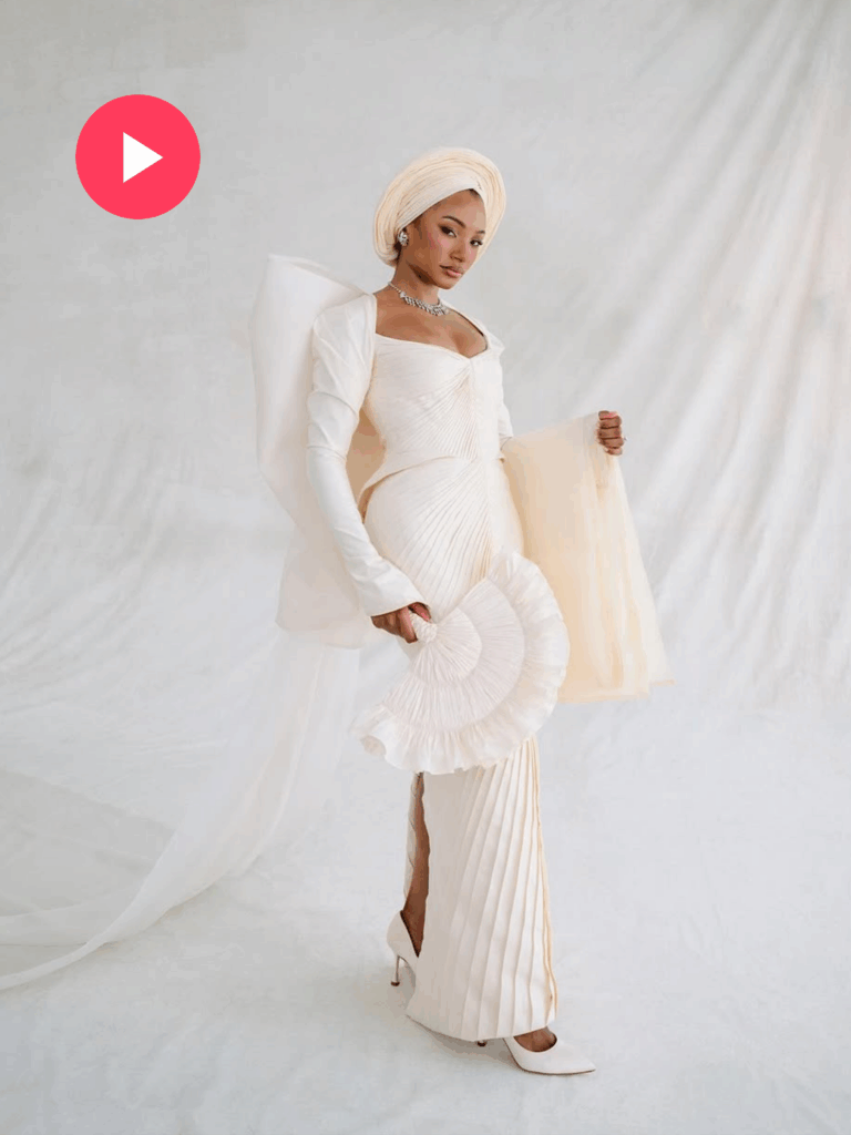 Temi Ajibade dressed in a fitted off-white gown with dramatic sleeves and a gele headwrap holds a matching hand fan while posing in front of a soft white draped background.