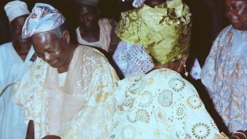 Elderly Nigerian couple in traditional attire dancing joyfully at a celebration, surrounded by others in colorful Yoruba lace and agbada outfits.